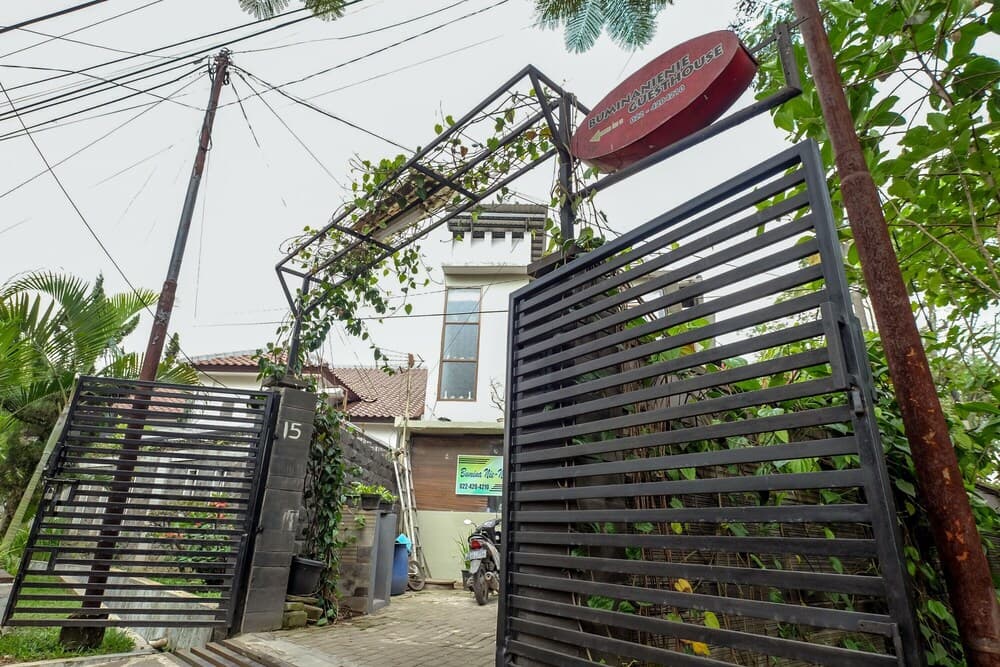 A tranquil tropical guesthouse entrance features a metal gate, lush greenery, and a red sign, ideal for travelers seeking a serene accommodation experience.
