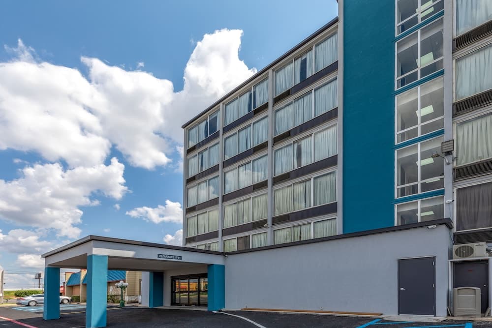A modern motel with a blue and gray facade stands under a bright sky with fluffy clouds, featuring a covered entrance and ample parking, ideal for travelers seeking convenient accommodations.