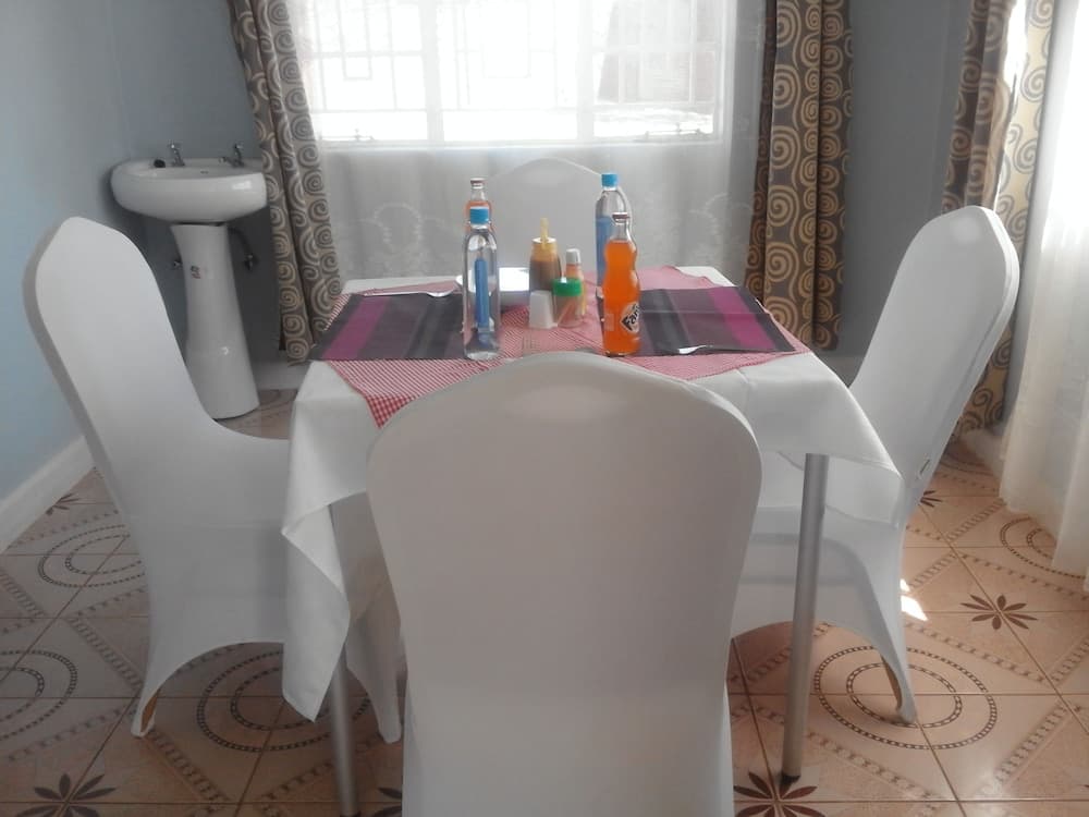 A cozy dining area with white chairs around a table set with colorful condiments and drinks, featuring patterned curtains and tiled flooring, ideal for travelers seeking a homely and inviting accommodation setting.