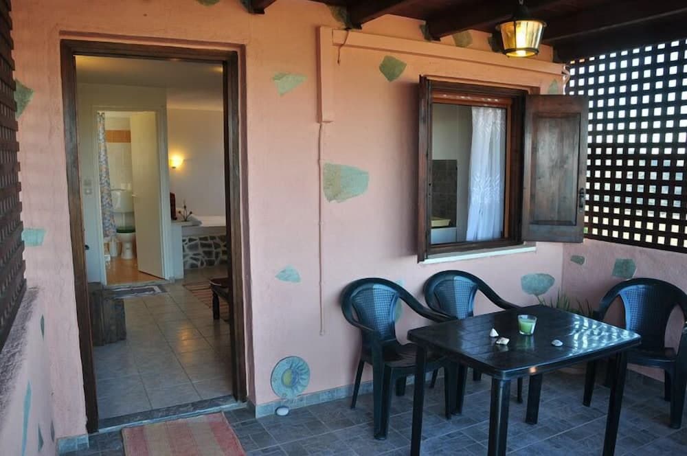 A cozy outdoor patio with a pink wall adorned with artistic patches, featuring a small dining set under a wooden ceiling light, an open door leading to a bathroom, and a lattice window, ideal for travelers seeking a quaint and intimate accommodation experience.