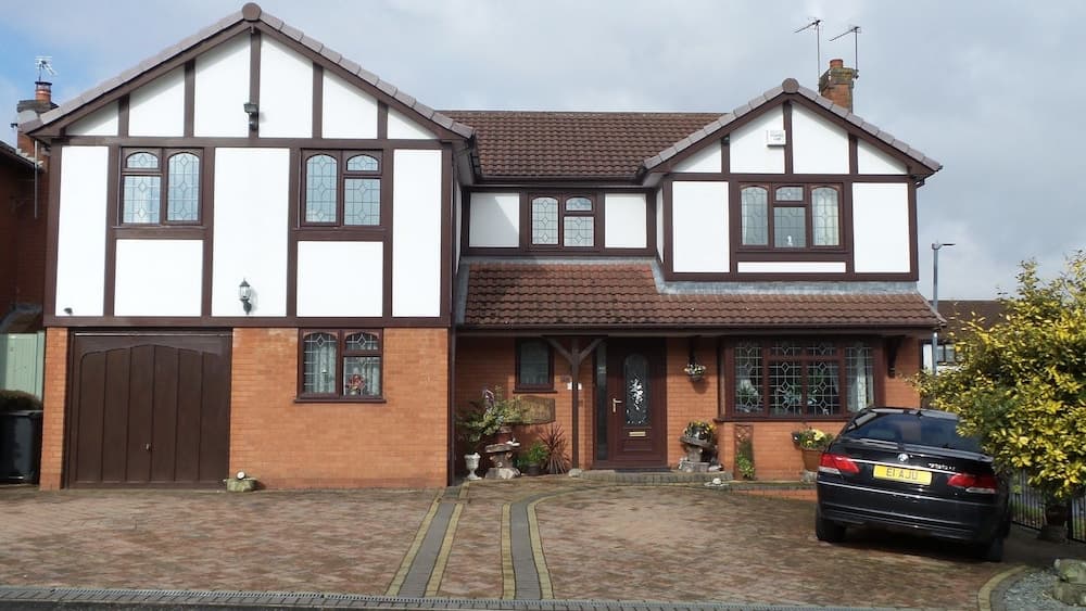 A charming Tudor-style house with white walls, brown timber framing, and a brick driveway exudes suburban comfort, featuring stained glass windows, potted plants, and a parked car, ideal for family-oriented travelers seeking a cozy residential retreat.