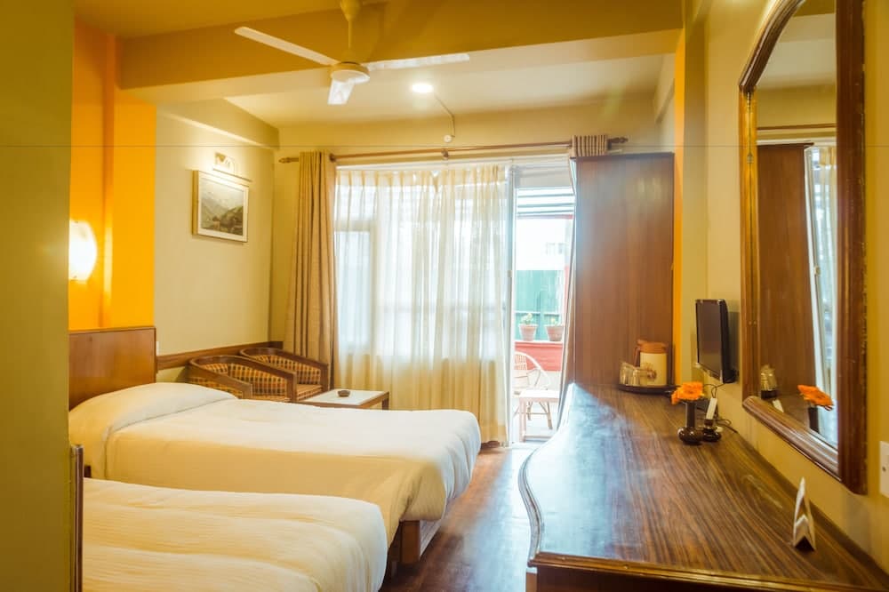 A cozy hotel room with warm yellow walls, two neatly made beds, a wooden vanity with a mirror, and a balcony view through sheer curtains, ideal for travelers seeking comfort and relaxation.