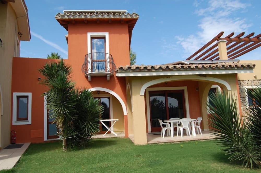 A vibrant orange villa with arched doorways, a balcony, and lush greenery offers a serene Mediterranean retreat perfect for family vacations.