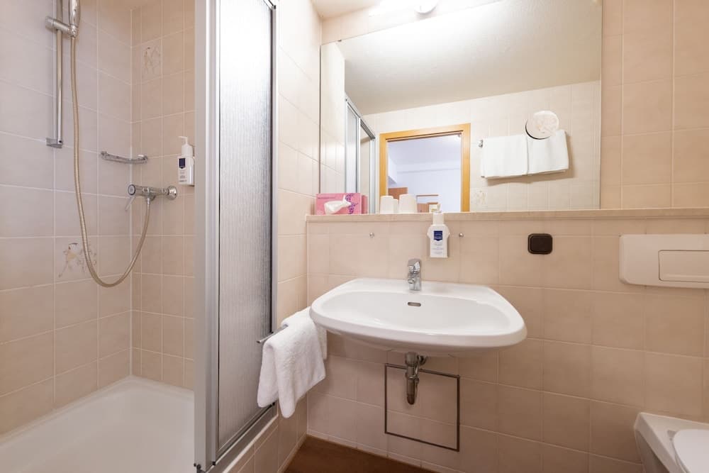 A cozy hotel bathroom features beige tiled walls, a bathtub with a shower, a sink with toiletries, and a mirror reflecting a warm ambiance, ideal for travelers seeking comfort and cleanliness.
