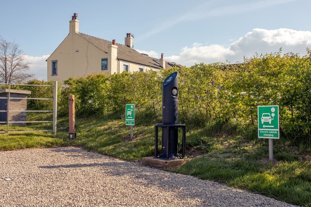 A serene rural roadside features an electric vehicle charging station amidst lush greenery, with a quaint yellow house in the background, ideal for eco-friendly travelers seeking a peaceful stopover.