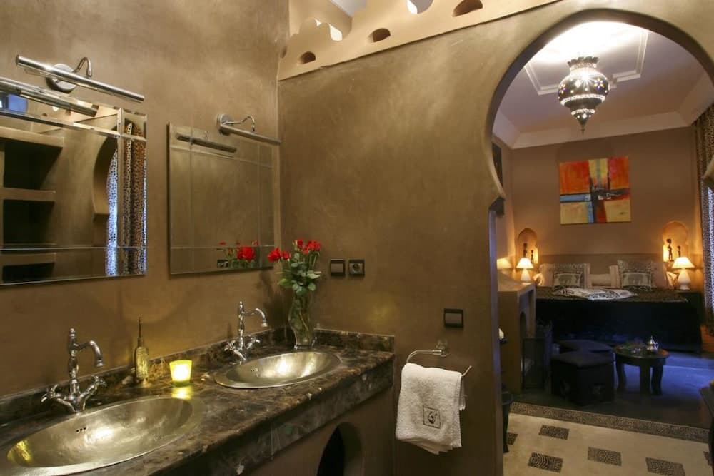 A luxurious Moroccan-style bathroom with dual sinks, warm earthy tones, and an arched doorway leading to a cozy bedroom adorned with vibrant art and ornate lighting, ideal for travelers seeking a serene and exotic retreat.