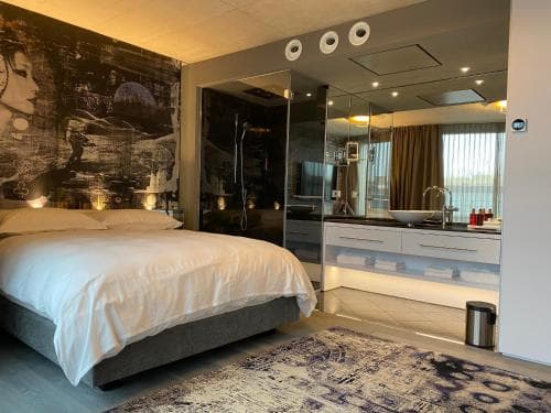 A modern hotel suite features a cozy bed adorned with white linens, a striking wall mural, a sleek bathroom with a glass shower, and a stylish vanity area, creating an inviting atmosphere perfect for travelers seeking comfort and contemporary design.