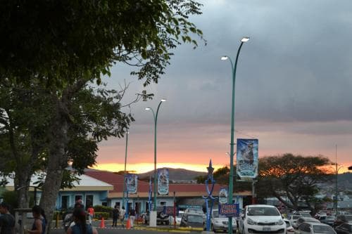A serene twilight scene at a bustling town square features silhouetted trees, vibrant streetlights, and colorful banners, evoking a peaceful yet lively atmosphere ideal for travelers exploring local culture.