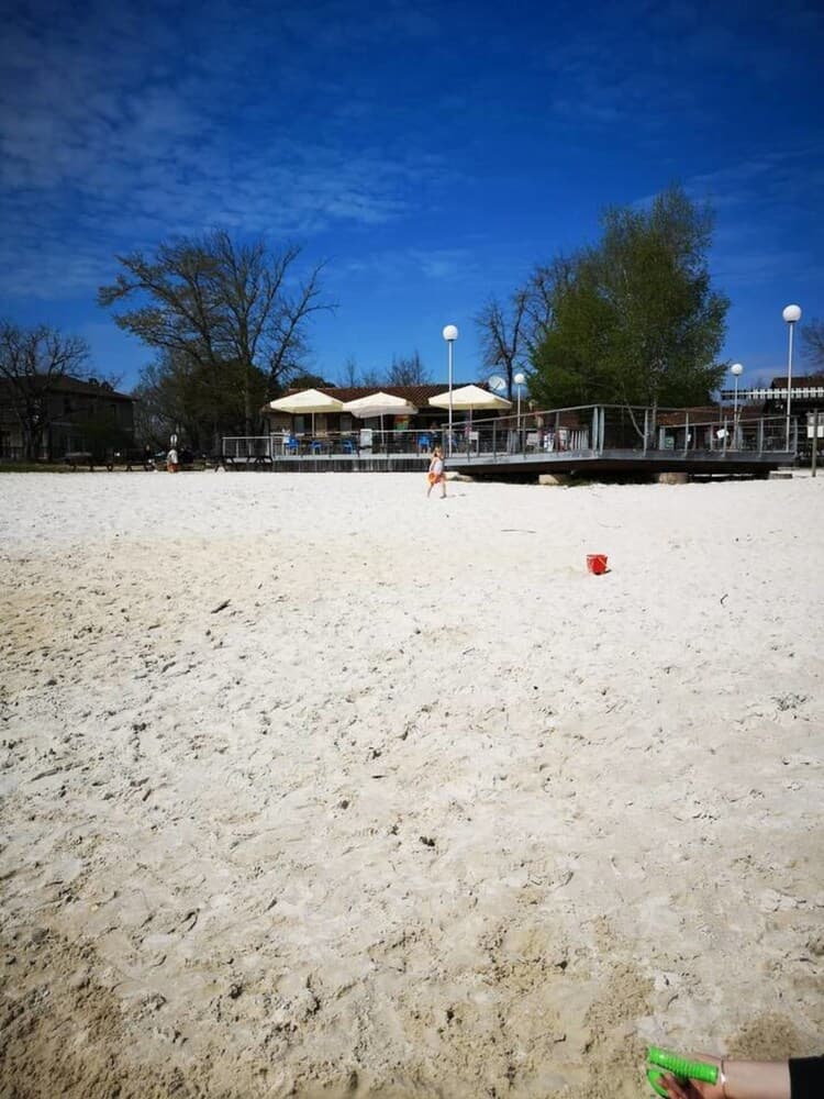 A serene sandy beach under a vibrant blue sky features a child playing near colorful buckets, with a relaxed seaside café and lush greenery in the background, perfect for family-friendly beach outings.