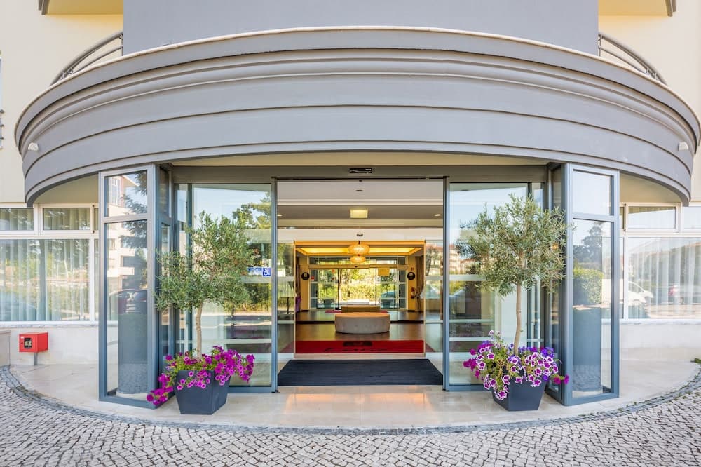 A modern hotel entrance features sleek glass doors framed by curved architecture, vibrant purple flowers, and olive trees, inviting travelers to a luxurious stay in a serene urban setting.