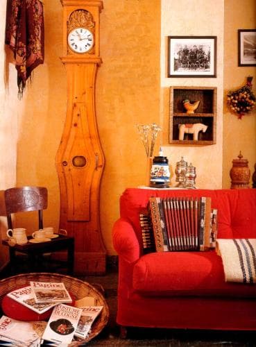A cozy living room with warm yellow walls, a red sofa adorned with an accordion, a wooden grandfather clock, and rustic decor creates a nostalgic atmosphere perfect for a cultural heritage getaway.