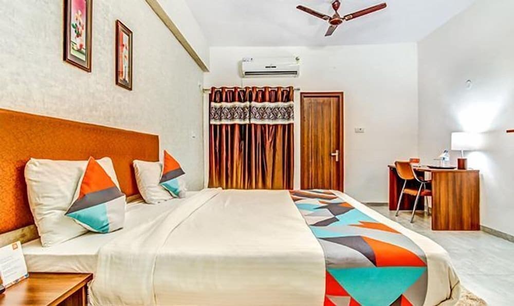 A cozy hotel room features a neatly made bed with colorful pillows, wooden furniture, patterned curtains, and a fan, ideal for travelers seeking comfort and convenience.