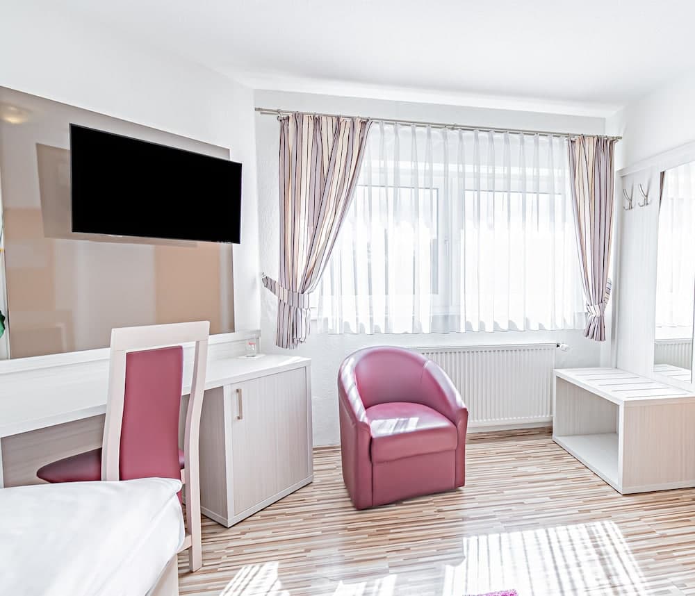 A cozy hotel room features a plush pink armchair by a bright window with sheer curtains, a sleek desk setup with a mounted TV, and modern furnishings, ideal for travelers seeking comfort and functionality.