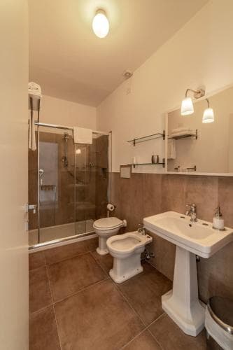 A cozy hotel bathroom features warm-toned tiled floors, a glass-enclosed shower, a pedestal sink, and soft lighting, creating an inviting atmosphere ideal for travelers seeking comfort and cleanliness.