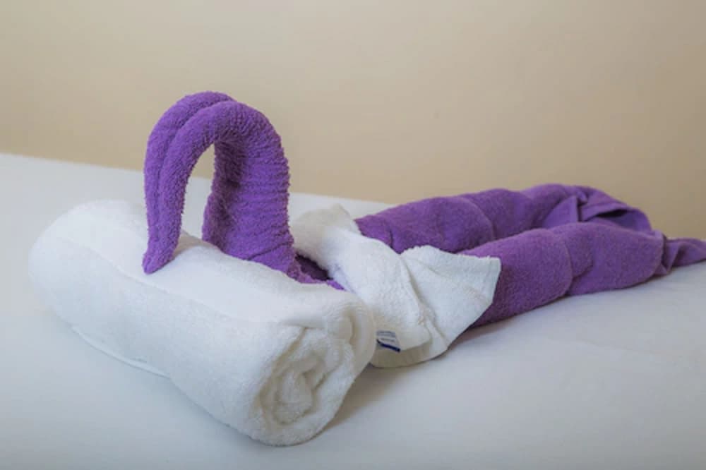 A serene hotel room scene features a creatively folded purple and white towel swan on a crisp white bed, evoking a sense of luxury and relaxation ideal for travelers seeking comfort and attention to detail.