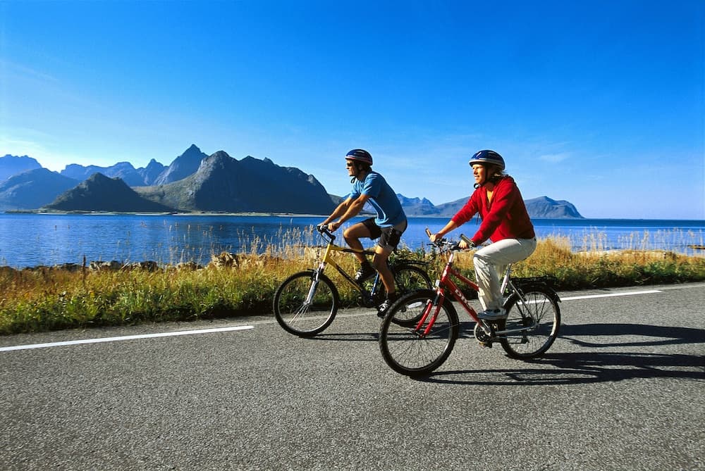 Two cyclists enjoy a scenic ride along a coastal road under a clear blue sky, with majestic mountains in the background, ideal for adventure travelers seeking outdoor activities.