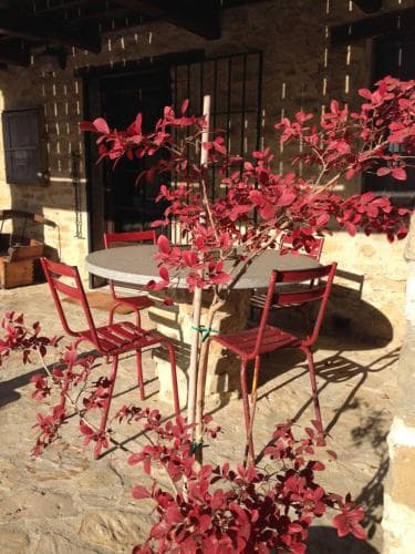A cozy outdoor patio features vibrant red chairs around a stone table, adorned with autumnal foliage, set against the rustic charm of a stone building, ideal for travelers seeking a serene retreat in a quaint countryside setting.