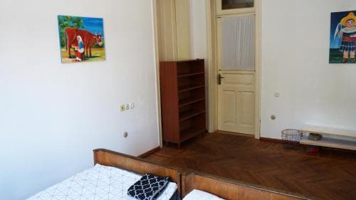 A cozy bedroom with wooden flooring, simple furnishings including a bed and bookshelf, adorned with colorful wall art, offers a comfortable and artistic retreat for travelers seeking a charming accommodation experience.