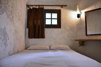 A cozy bedroom with rustic charm features a neatly made bed, a small window with patterned curtains, and a warm wall sconce, ideal for travelers seeking a quaint and intimate retreat.