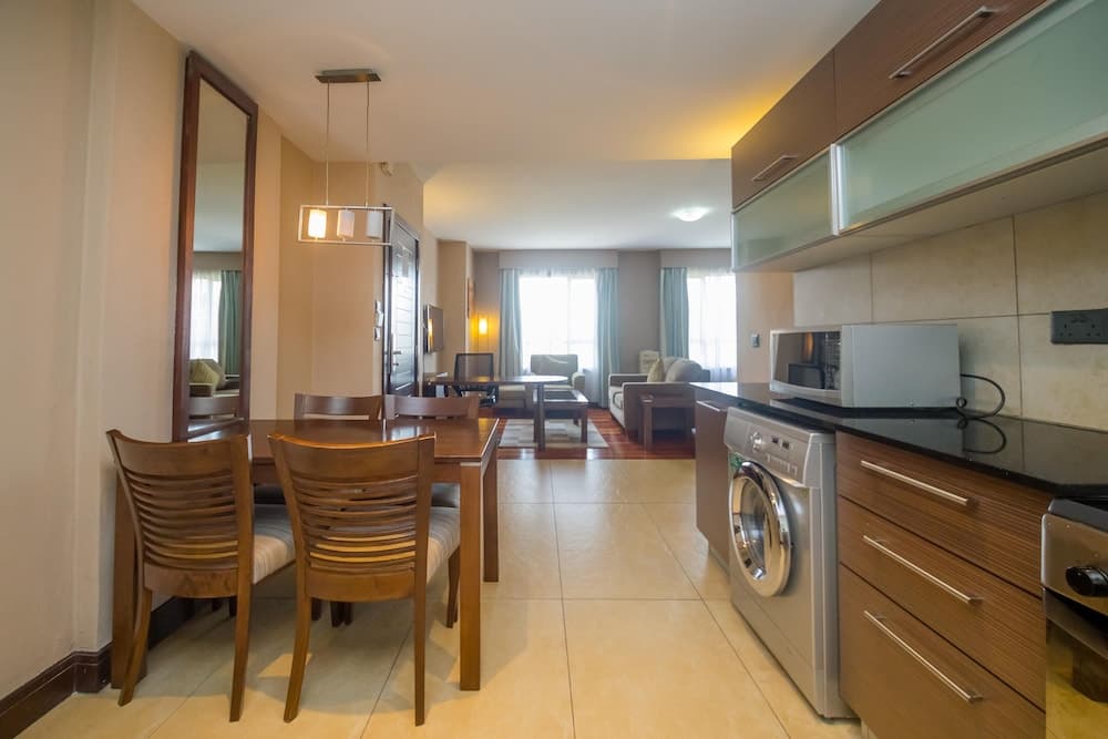 A cozy apartment interior features a well-lit dining area with wooden furniture, a modern kitchen with appliances, and a comfortable living space with large windows, ideal for travelers seeking a home-like stay.