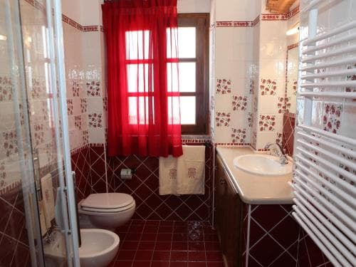 A cozy bathroom with red-tiled floors, floral-patterned walls, and a bright window adorned with red curtains offers a charming and inviting space for travelers seeking comfort in their accommodations.