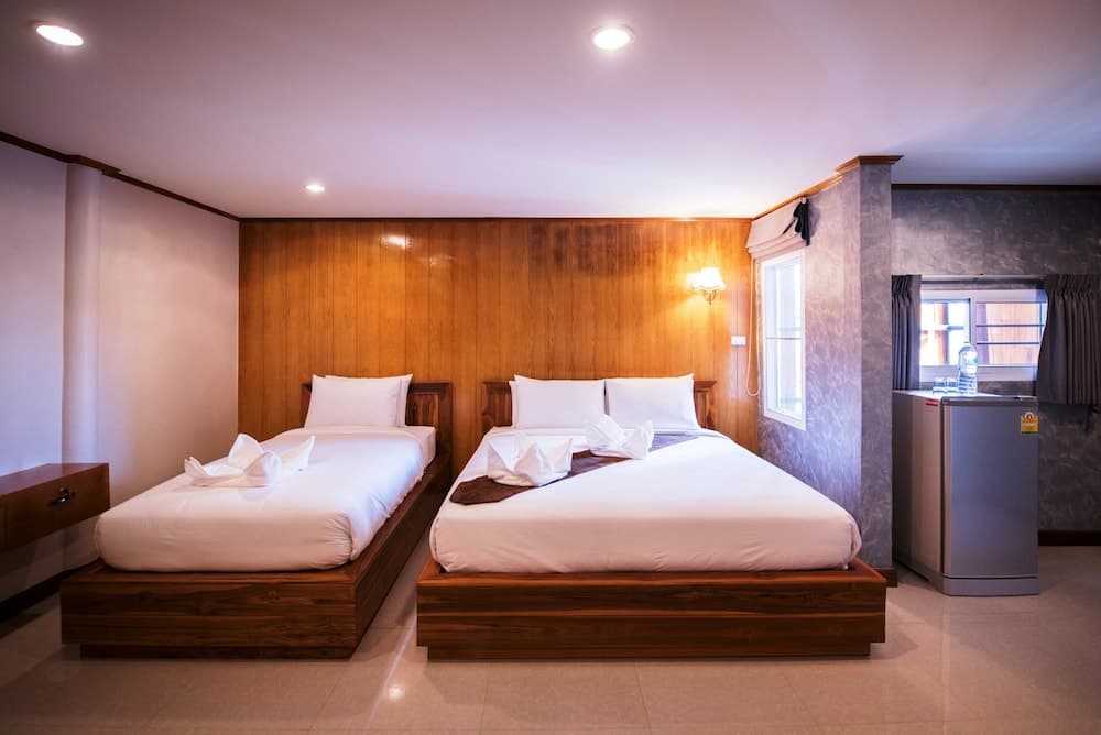 A cozy hotel room features two neatly made wooden beds with decorative towels, a small fridge by the window, and warm lighting, ideal for travelers seeking comfort and simplicity.