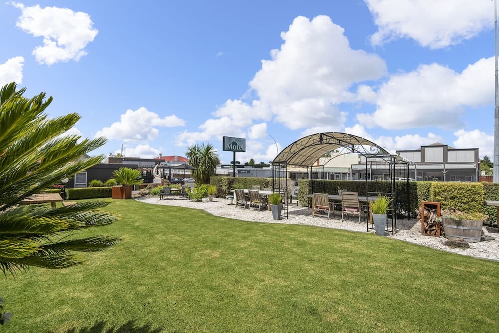 A serene motel courtyard features lush green lawns, cozy seating under a decorative arbor, and tropical plants, creating an inviting atmosphere ideal for travelers seeking relaxation.