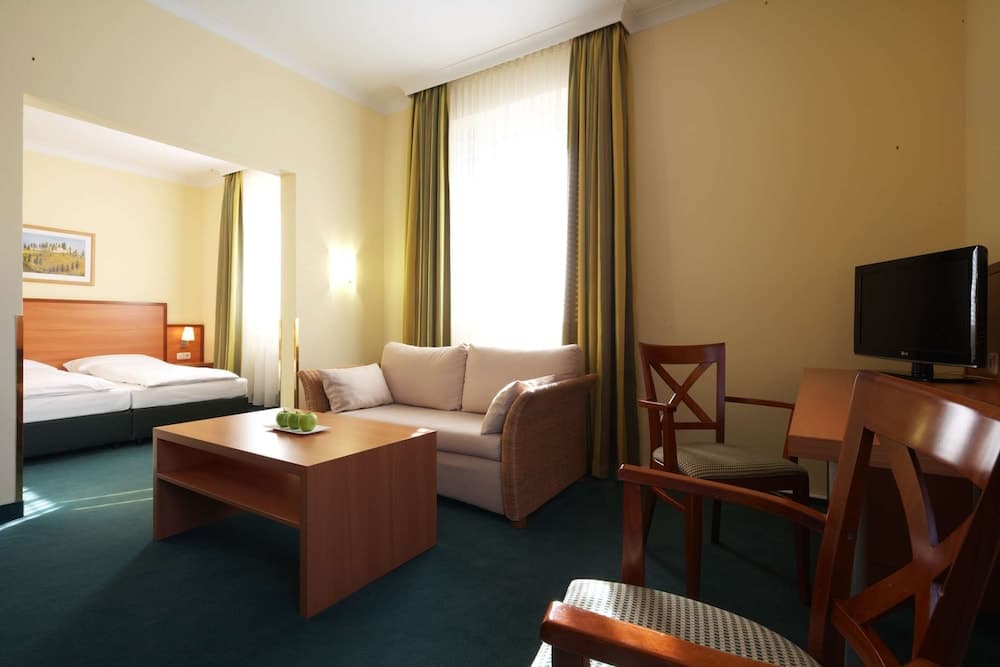 A cozy hotel room features a comfortable seating area with a sofa, wooden coffee table, and chairs near a desk with a TV, all bathed in soft light from large windows with striped curtains, ideal for travelers seeking relaxation and convenience.