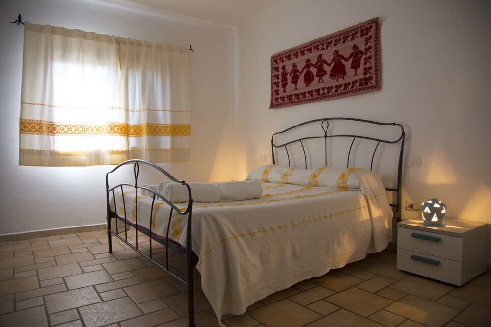 A cozy bedroom with a metal bedframe, tiled floor, embroidered curtains, and a cultural tapestry creates a warm and inviting atmosphere ideal for travelers seeking a comfortable and culturally rich stay.