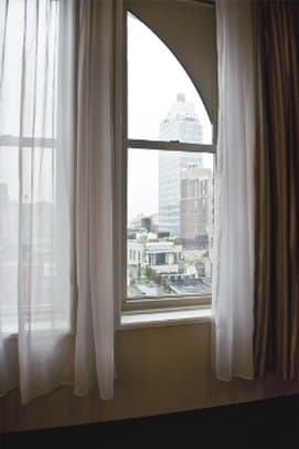 A serene hotel room window frames an urban skyline view through sheer curtains, ideal for travelers seeking cityscapes from cozy accommodations.