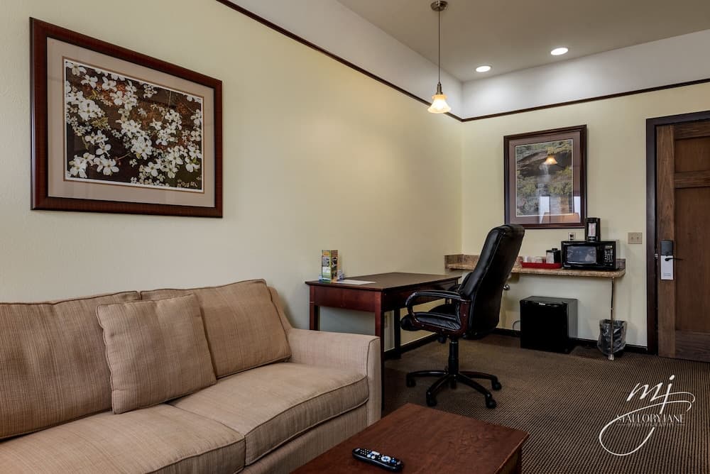 A cozy hotel suite features a beige sofa, wooden desk with an office chair, framed artwork on cream walls, and a small kitchenette area, ideal for travelers seeking comfort and functionality.