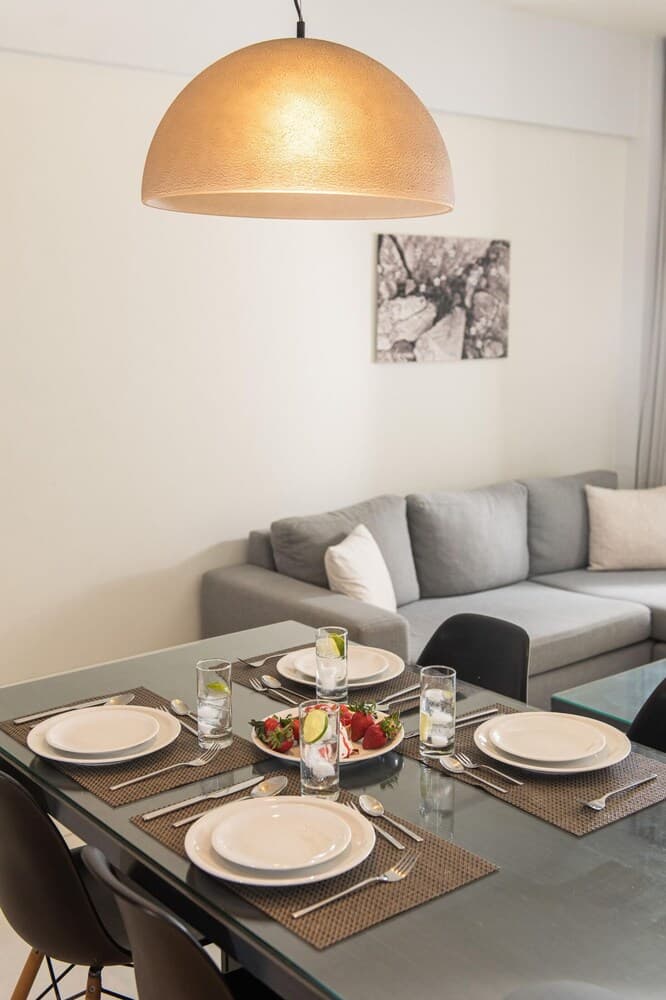 A cozy modern dining area features a sleek glass table set for four under a warm dome light, complemented by a plush gray sofa and minimalist decor, ideal for travelers seeking stylish apartment accommodations.