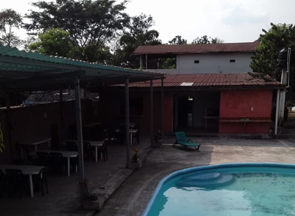A tranquil outdoor setting features a poolside area with lounge chairs, shaded dining tables under a green roof, and rustic buildings surrounded by lush greenery, ideal for a relaxing getaway or family vacation.