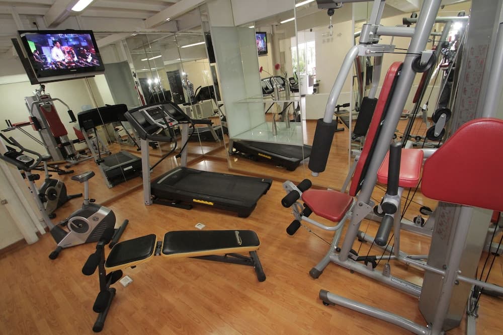 A well-equipped indoor gym features various fitness machines like treadmills and weight benches, surrounded by mirrors and a TV, creating an inviting atmosphere for travelers seeking a workout space during their stay.