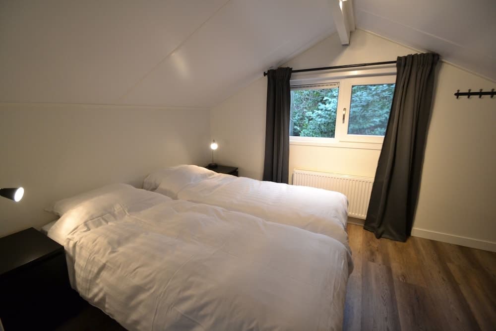 A cozy attic bedroom with sloped ceilings, twin beds dressed in white linens, wooden flooring, and a window offering a serene view of greenery, ideal for travelers seeking a tranquil retreat.