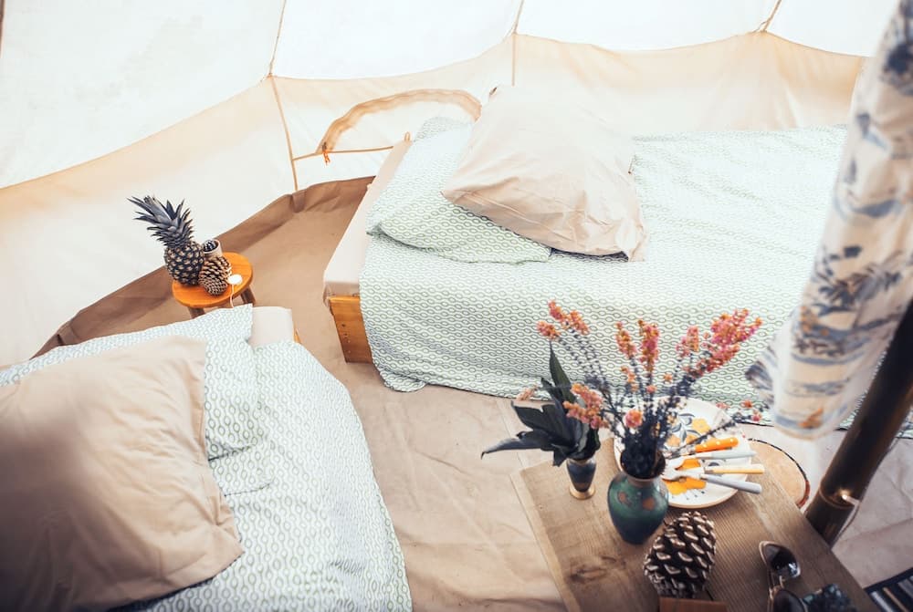 A cozy glamping tent interior features two beds adorned with green patterned linens, a rustic wooden table with flowers and pinecones, creating a serene and inviting atmosphere perfect for outdoor enthusiasts seeking comfort in nature.