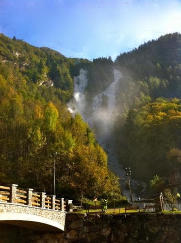A serene mountain valley features a majestic waterfall cascading through lush greenery, framed by a quaint stone bridge and wooden railing, ideal for tranquil nature retreats and scenic photography.