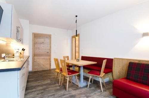 A cozy apartment interior features a modern kitchenette, wooden dining set, and red upholstered seating, creating a warm and inviting atmosphere ideal for travelers seeking comfort in a compact living space.