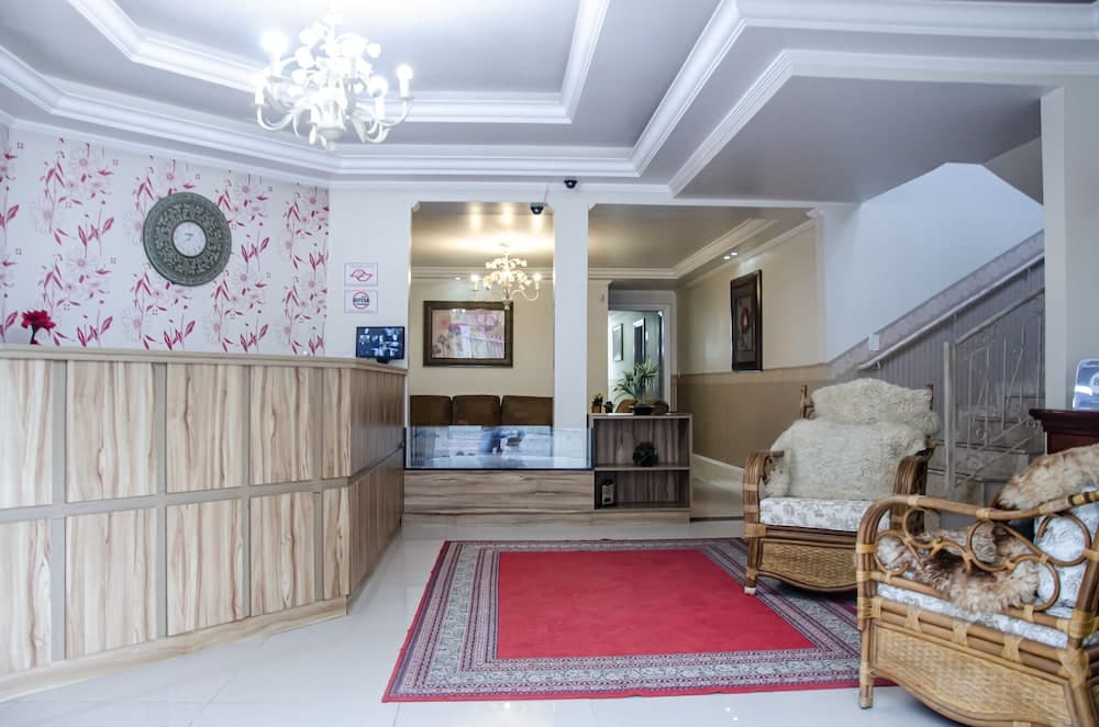 A cozy hotel lobby features a red patterned rug, wooden furniture draped with plush white throws, a sleek reception desk, and elegant chandeliers, creating a warm and inviting atmosphere ideal for travelers seeking comfort and style.