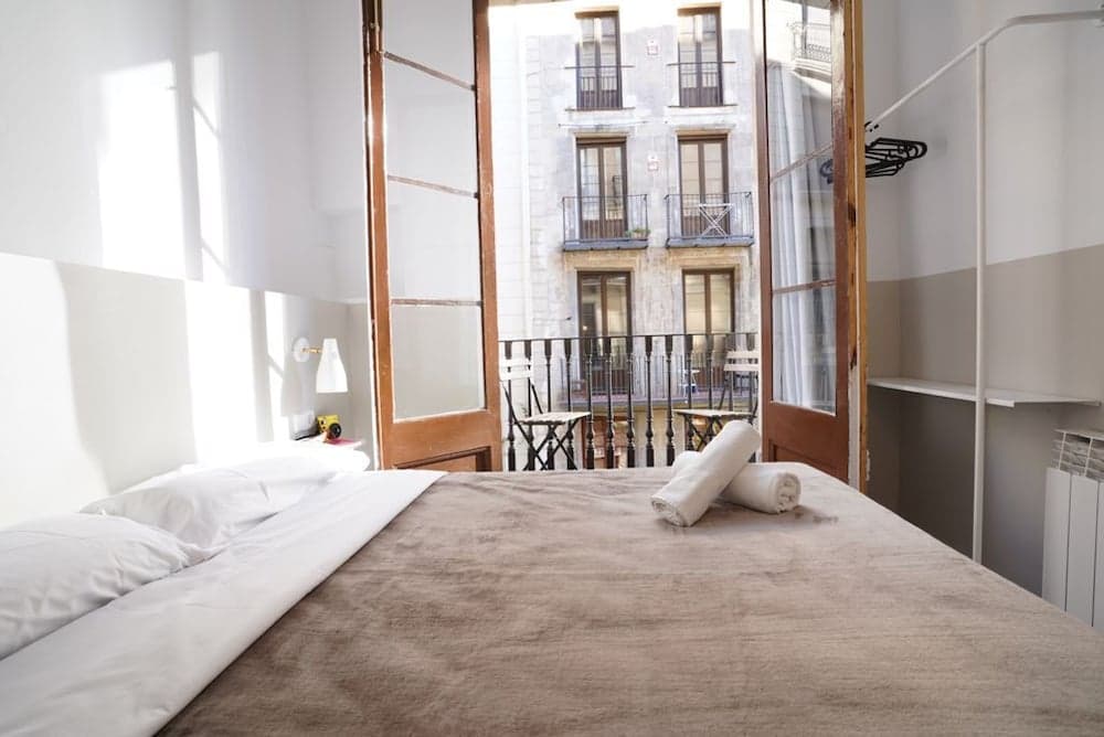 A cozy bedroom with a neatly made bed opens to a charming balcony overlooking a European-style building, ideal for travelers seeking a serene urban retreat.