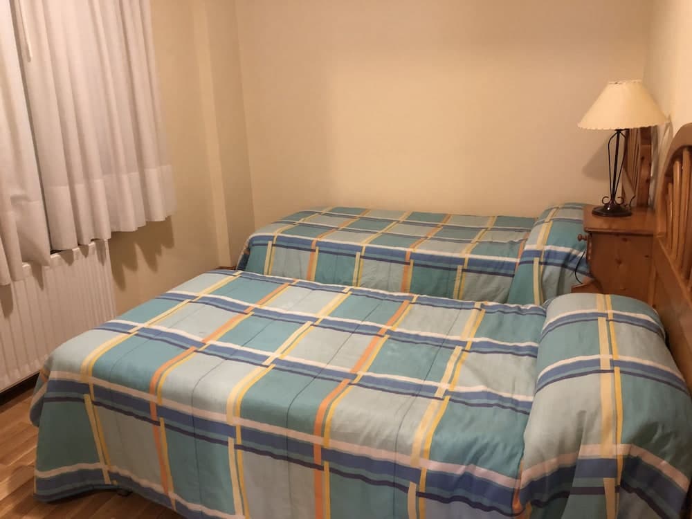 A cozy hotel room features two neatly made beds with blue plaid bedding, a wooden bedside table with a lamp, and soft curtains over a window, ideal for travelers seeking comfort and simplicity.