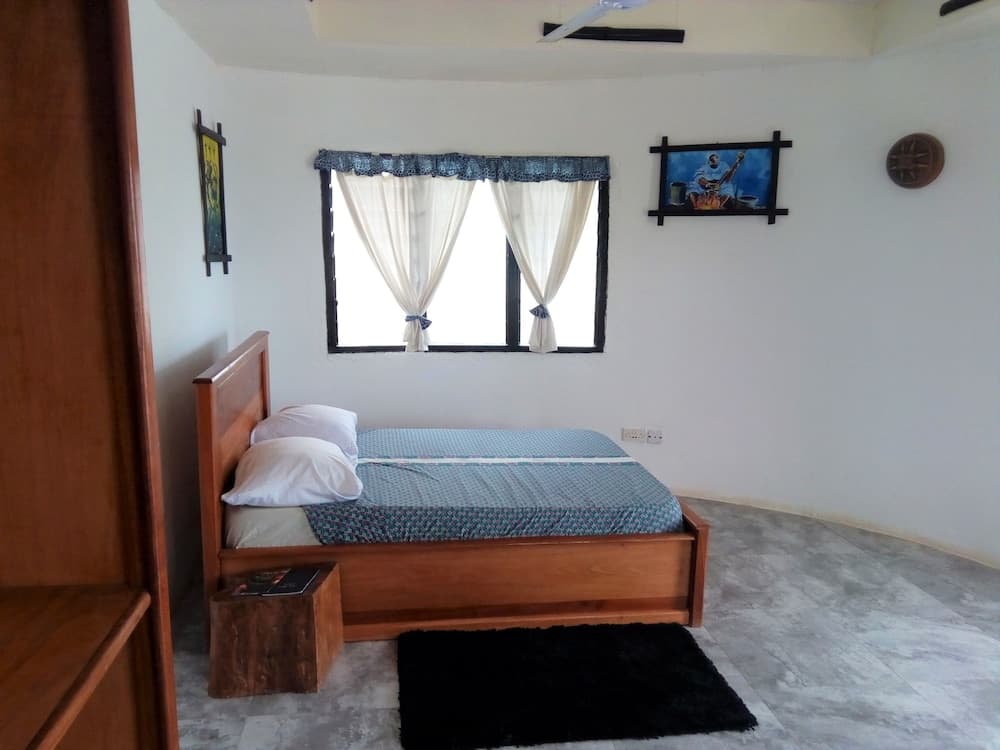 A cozy bedroom with a wooden bed, blue patterned bedding, sheer curtains over a window, and cultural wall art creates a serene atmosphere ideal for travelers seeking a peaceful retreat.