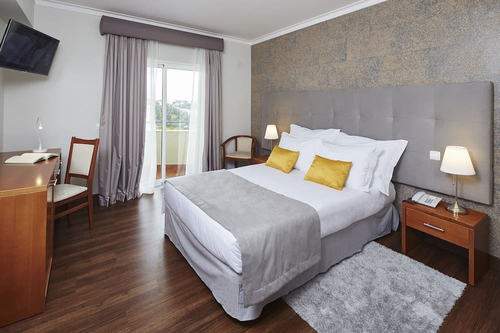 A cozy hotel room features a neatly made bed with golden pillows, wooden furnishings, a plush rug, and a balcony view, ideal for travelers seeking comfort and relaxation.