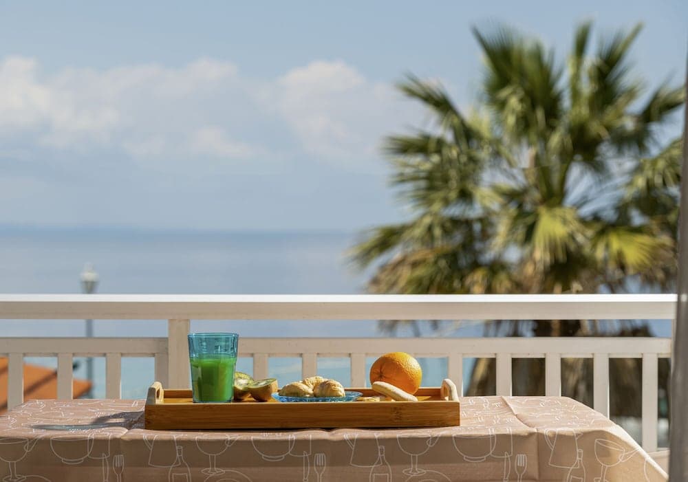 A serene coastal balcony scene features a breakfast tray with fresh juice and fruit, set against a backdrop of palm trees and the ocean, ideal for promoting luxurious seaside retreats.