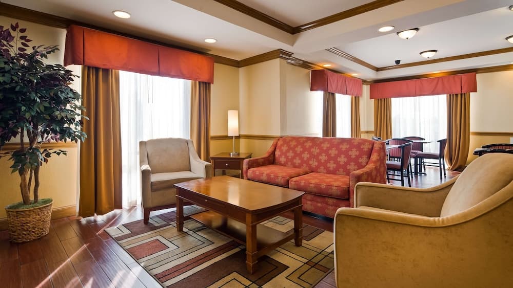 A cozy hotel lounge features warm-toned furniture, patterned rugs, and large windows with red valances, creating an inviting atmosphere ideal for travelers seeking relaxation or business meetings.