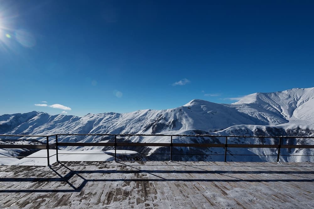 A serene winter vista from a wooden observation deck overlooks snow-covered mountains under a clear blue sky, ideal for adventure seekers planning a ski or hiking trip.