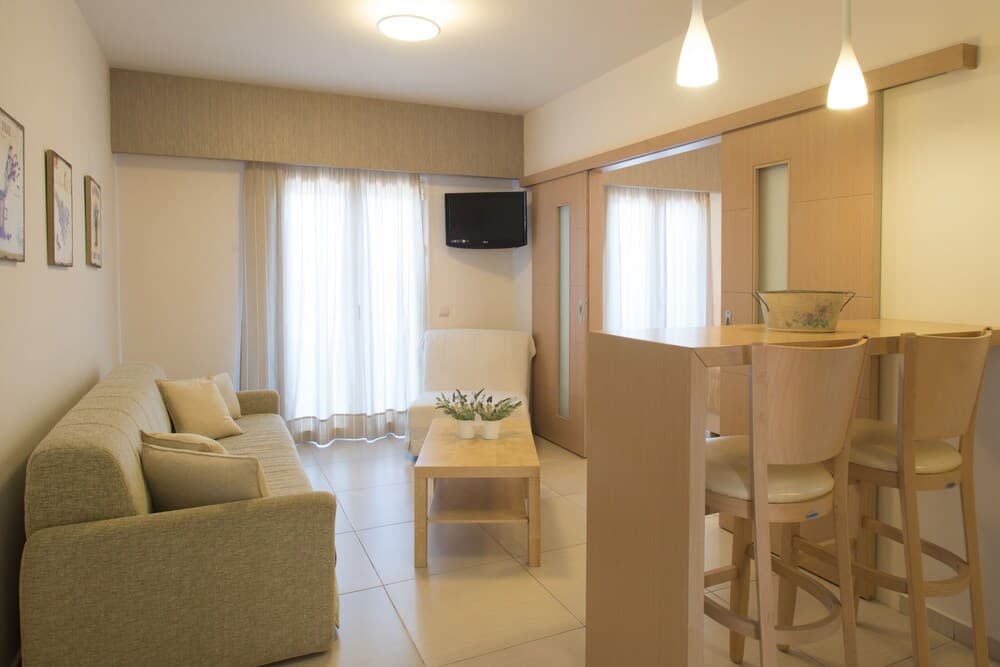 A cozy apartment living space features a comfortable sofa, wooden dining set, soft lighting, and large windows draped in sheer curtains, ideal for travelers seeking a warm and inviting accommodation.