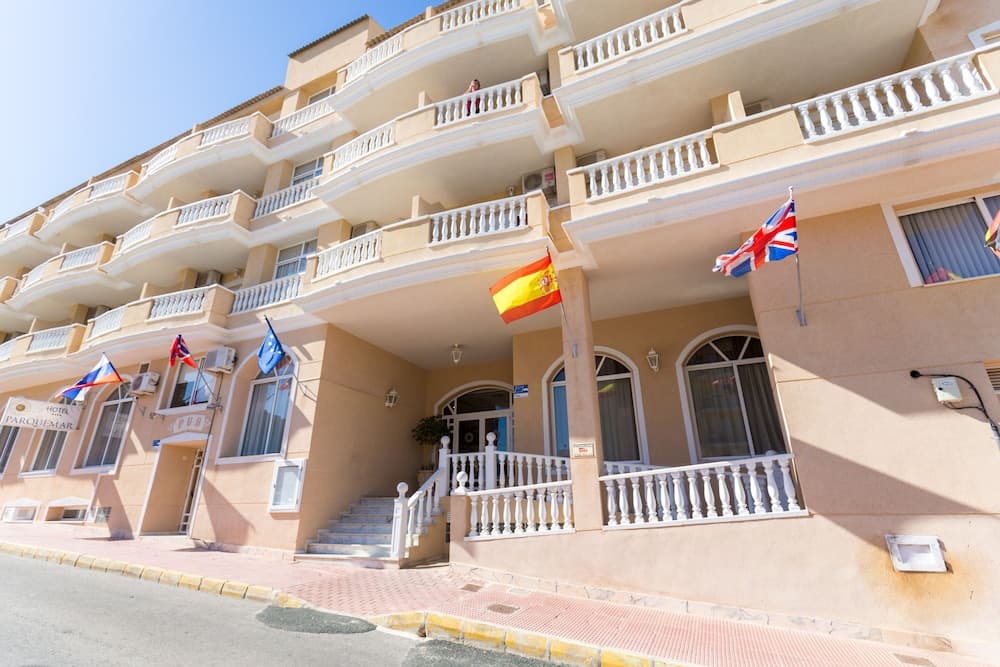 A sunny day highlights the inviting facade of a multi-story hotel adorned with Spanish and British flags, featuring balconies and a welcoming entrance, ideal for travelers seeking a cozy stay in a vibrant coastal town.