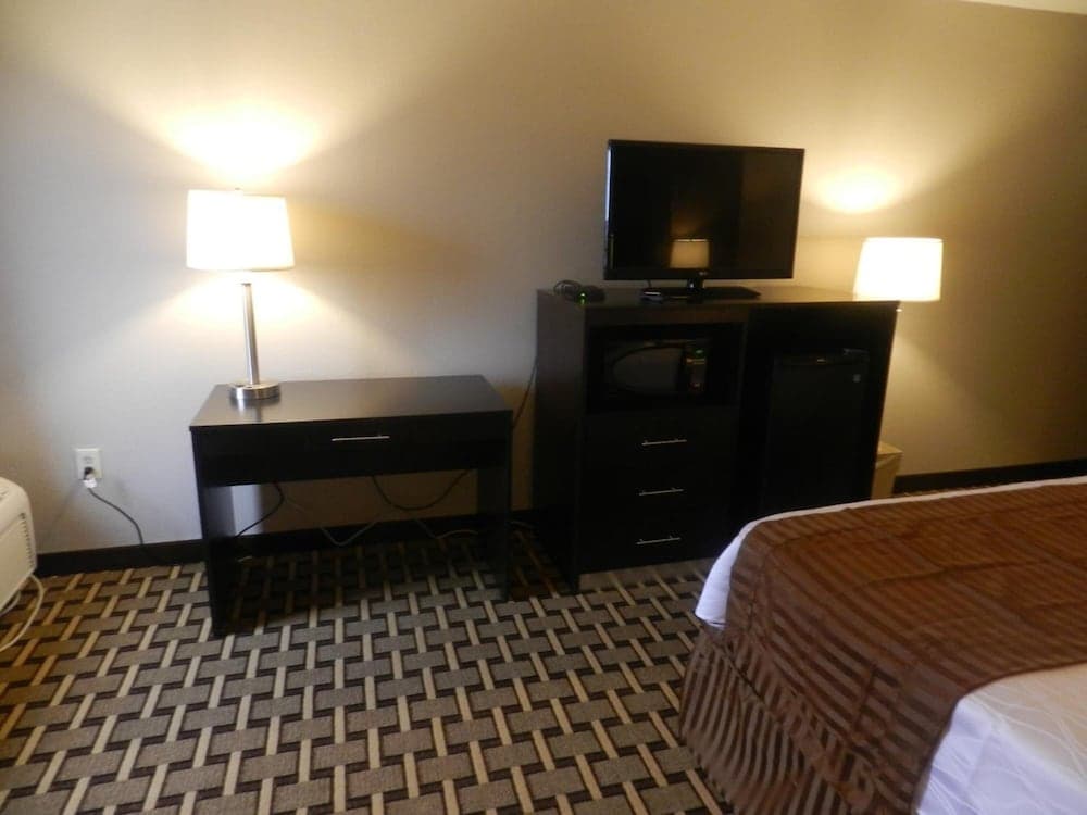 A cozy hotel room features a neatly made bed, a desk with a lamp, a TV atop a cabinet with a microwave and mini-fridge, all set against warm lighting, ideal for travelers seeking comfort and convenience.