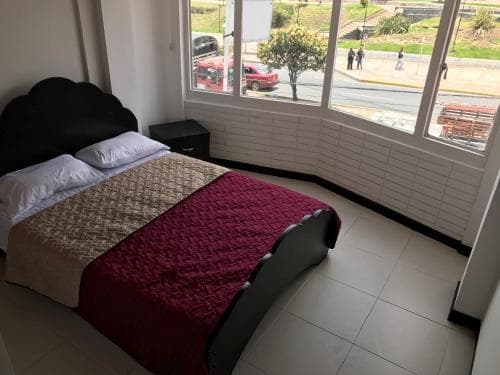 A cozy hotel room with a neatly made bed adorned in red and beige bedding overlooks a lively street scene through large windows, ideal for travelers seeking comfort and convenience.